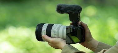 Usage image showing left-side view of camera hand-held by user, with microphone and wind screen attached