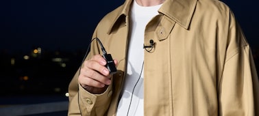 Usage image of a man in an outdoor setting holding an ECM-W3 transmitter connected to an ECM-L1 microphone with metal windscreen on his jacket lapel