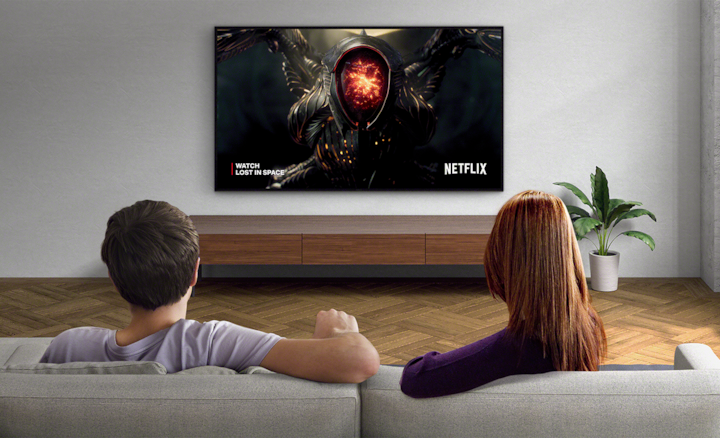 Two people sitting on a sofa watching a wall-mounted TV with screenshot of Lost in Space and Netflix logo bottom right