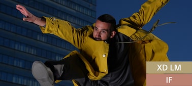 Against the backdrop of urban buildings, a short-haired man wearing a yellow jacket is captured in the moment of jumping.