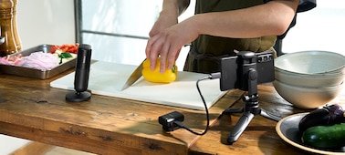 Usage image showing a person cooking with the microphone standing nearby, with the receiver connected to a smartphone on a stand via a USB cable