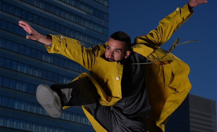 Against the backdrop of urban buildings, a short-haired man wearing a yellow jacket is captured in the moment of jumping.