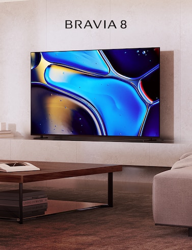 Living room scene with light brown sofa to right, coffee table center with book, and BRAVIA 8 TV on a plinth with ornaments to left and screenshot of blue water droplets