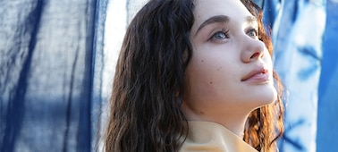 A portrait of a woman looking upwards and gazing into the distance. The focus is on her right eye and hair, showcasing the high resolution. The blue cloths in the background are smoothly blurred